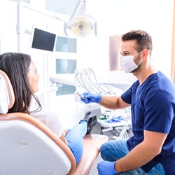 A woman seated in a dentist’s chair and smiling at her dentist as he prepares to check her teeth and gums