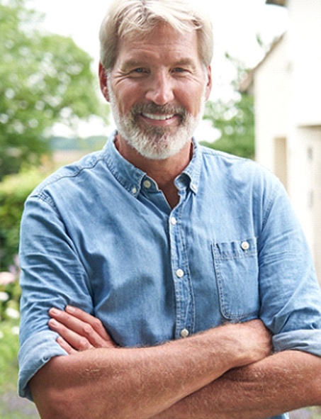 An older man wearing a denim button-down shirt and standing outside his home, smiling after learning about the cost of dental implants in Myrtle Beach