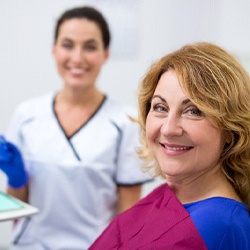 A dental professional smiling in the background while a middle-aged female patient smiles in preparation for her dental implant consultation