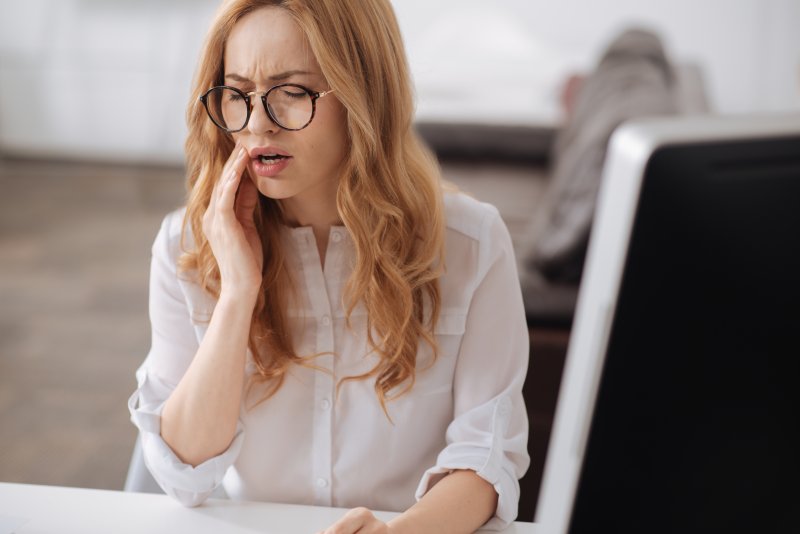 a young woman wearing glasses pressing her hand to her cheek because of pain she’s experiencing